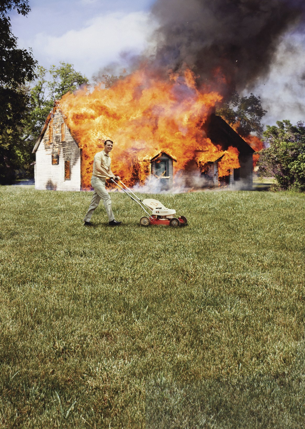 Man mowing a lawn in front of a burning house